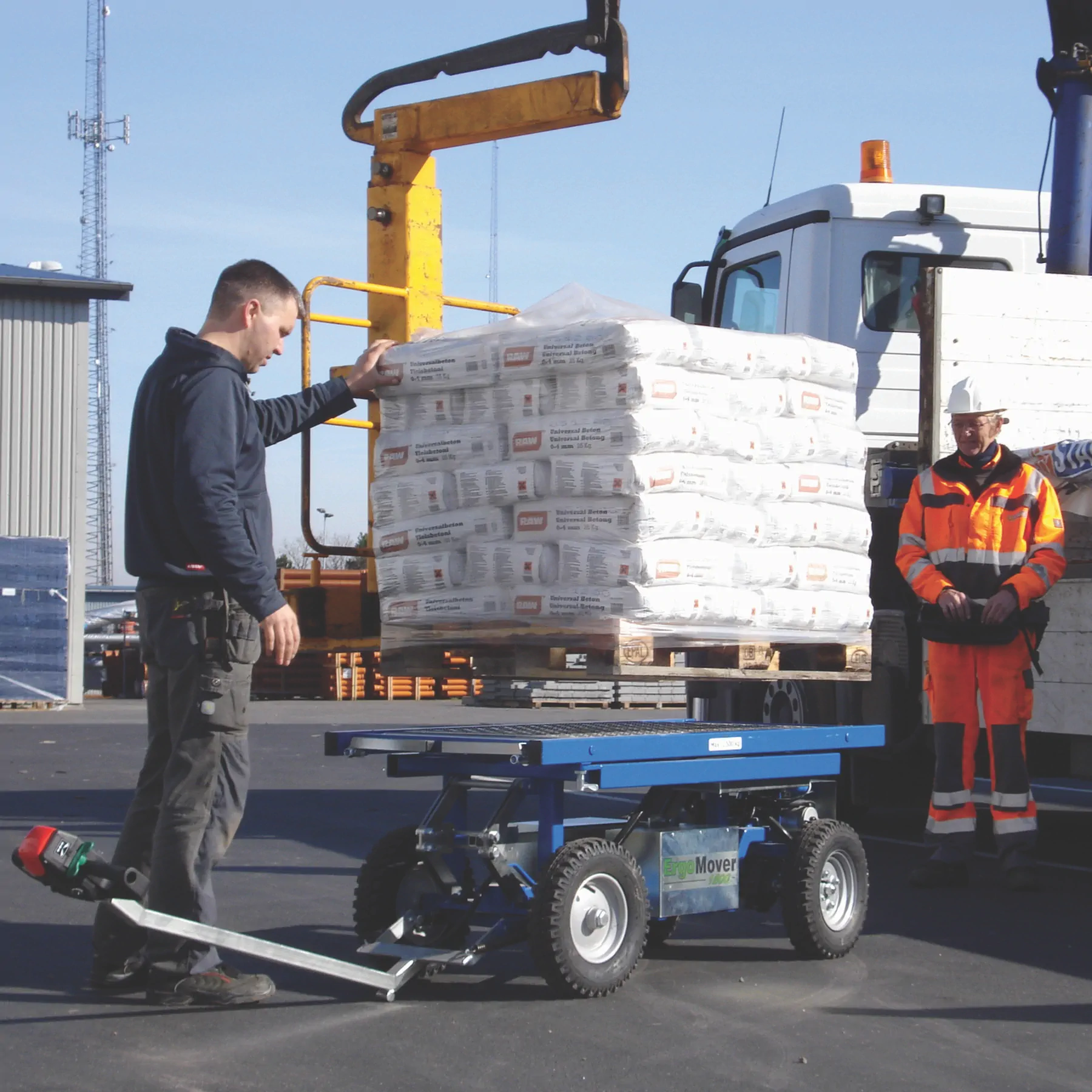 Worker steering a Lift and Go ErgoMover with a low blue deck beside a pallet of bagged material and a delivery truck.