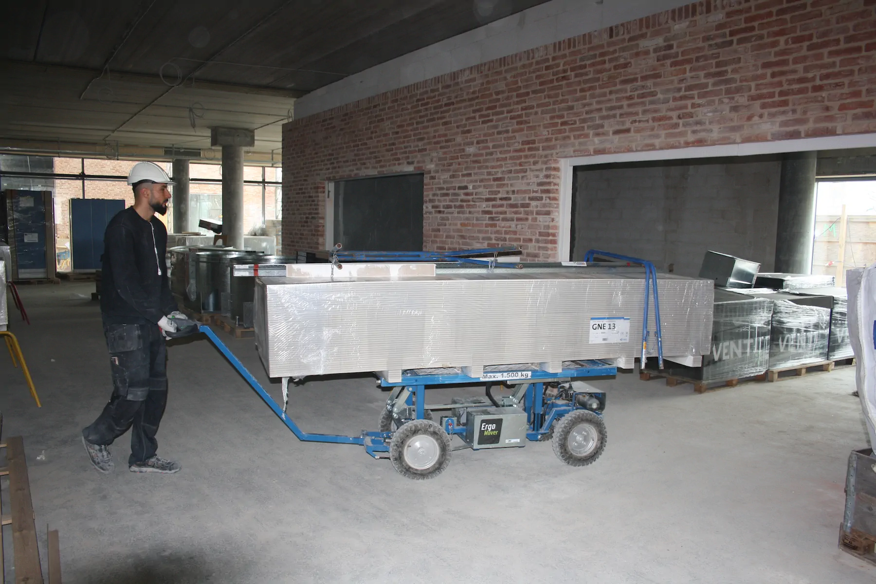 Worker in a white hard hat walking behind a drawbar ErgoMover that carries a stack of plasterboard through an unfinished concrete building.