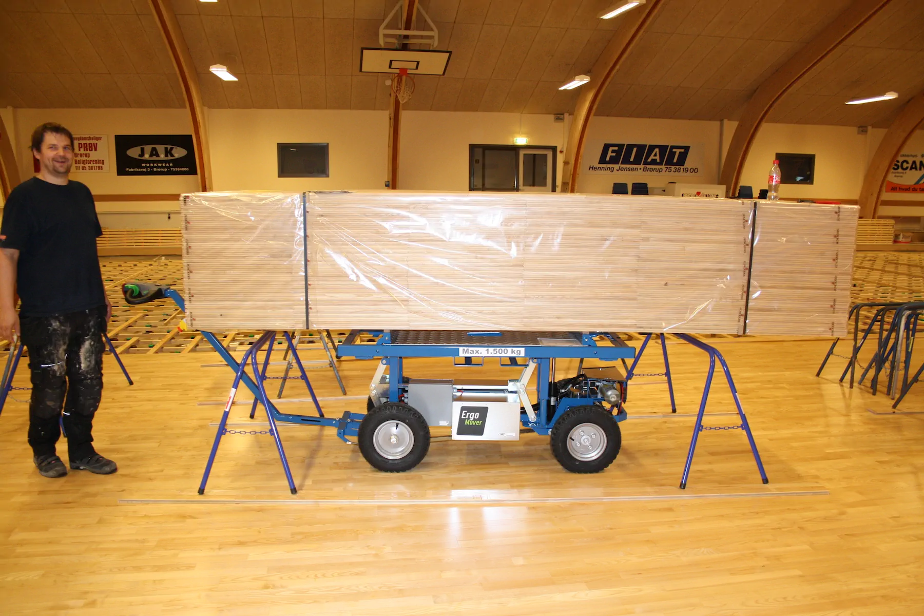Worker standing beside an ErgoMover electric cart that carries a long wrapped stack of timber boards on trestles in a sports hall.