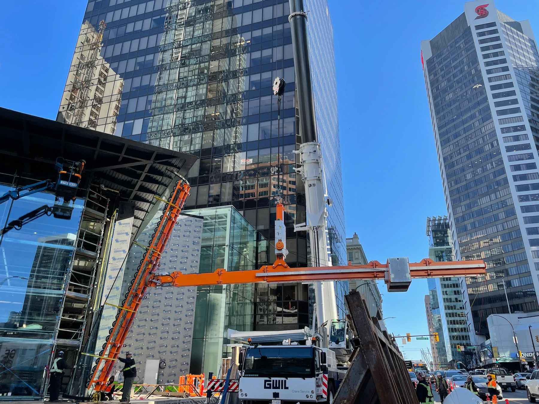Orange lifting beam hanging from a mobile crane beside a glass tower, with a long orange pad rail leaning against the facade at left and crew working below.