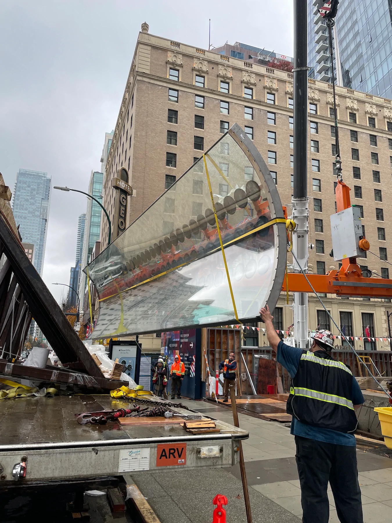 Curved glass pane tilted in the air above a flatbed trailer, held by an orange lifting arm on a crane hook while a worker guides it by hand.
