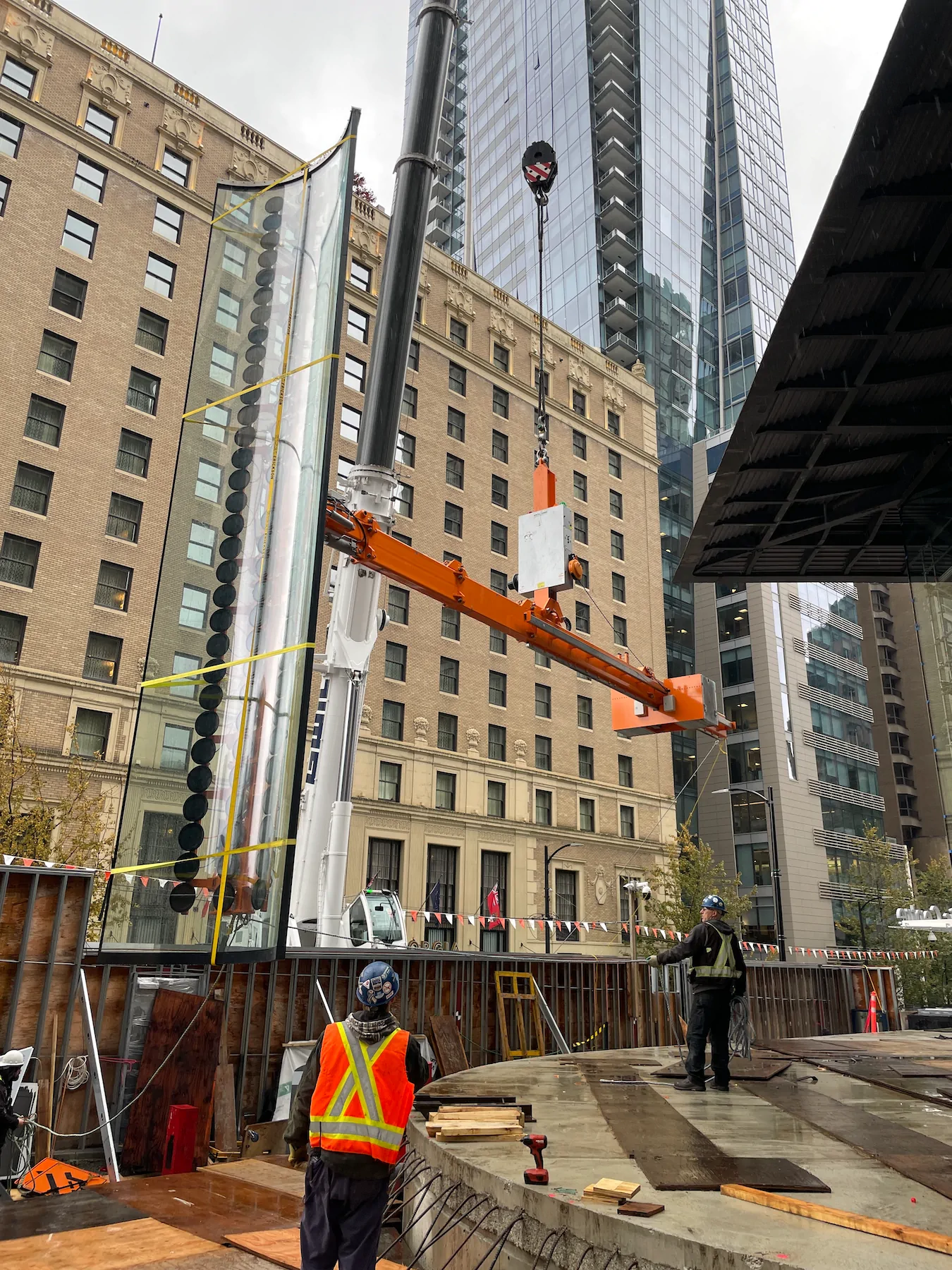 Tall curved glass pane held on a vertical pad rail beside an orange lifting arm and a white crane mast, between a brick hotel and a glass tower.
