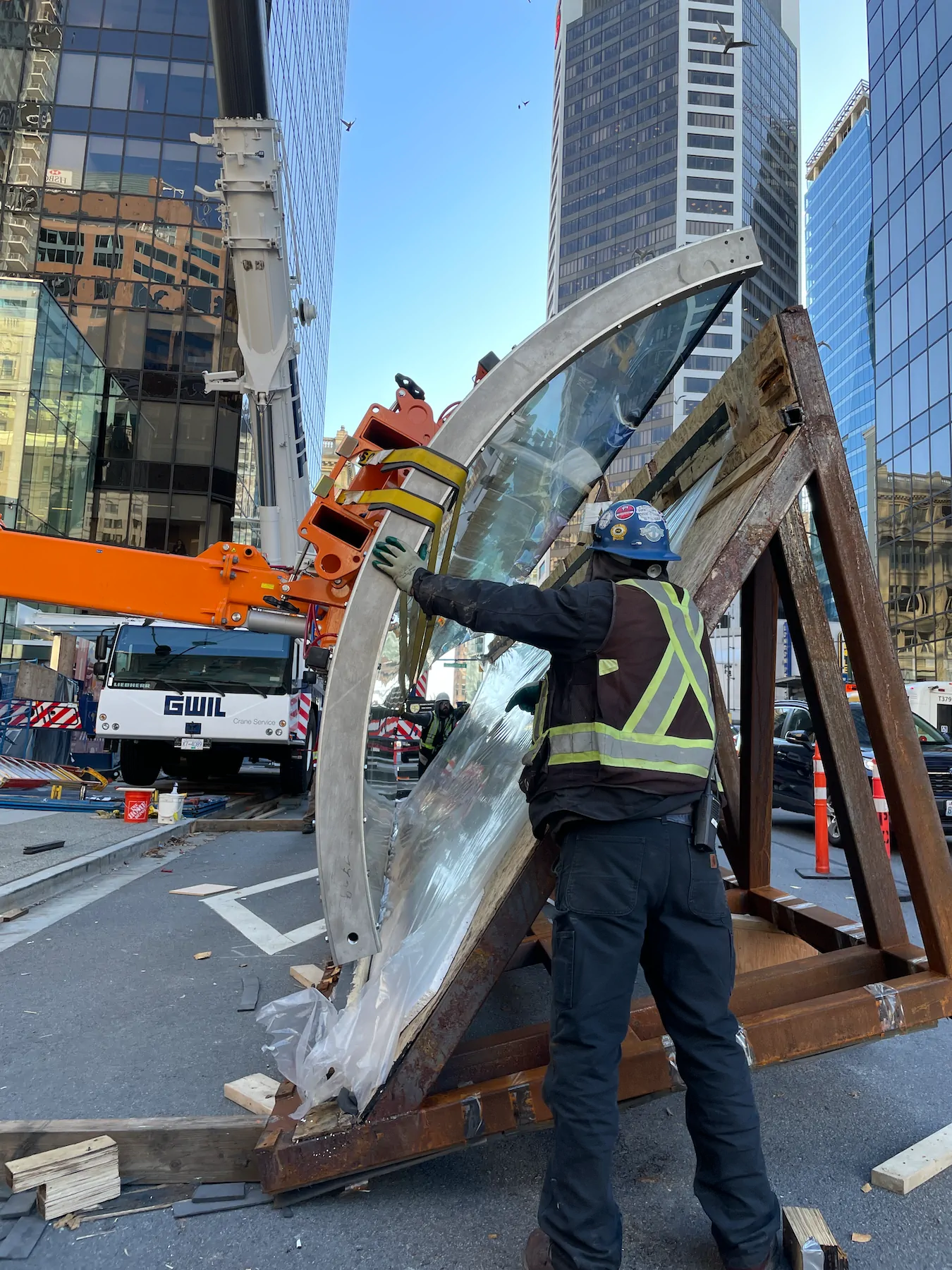 Worker in a hard hat and high-visibility vest steadying a curved glass pane in a steel rack, with an orange lifting arm and a mobile crane behind.