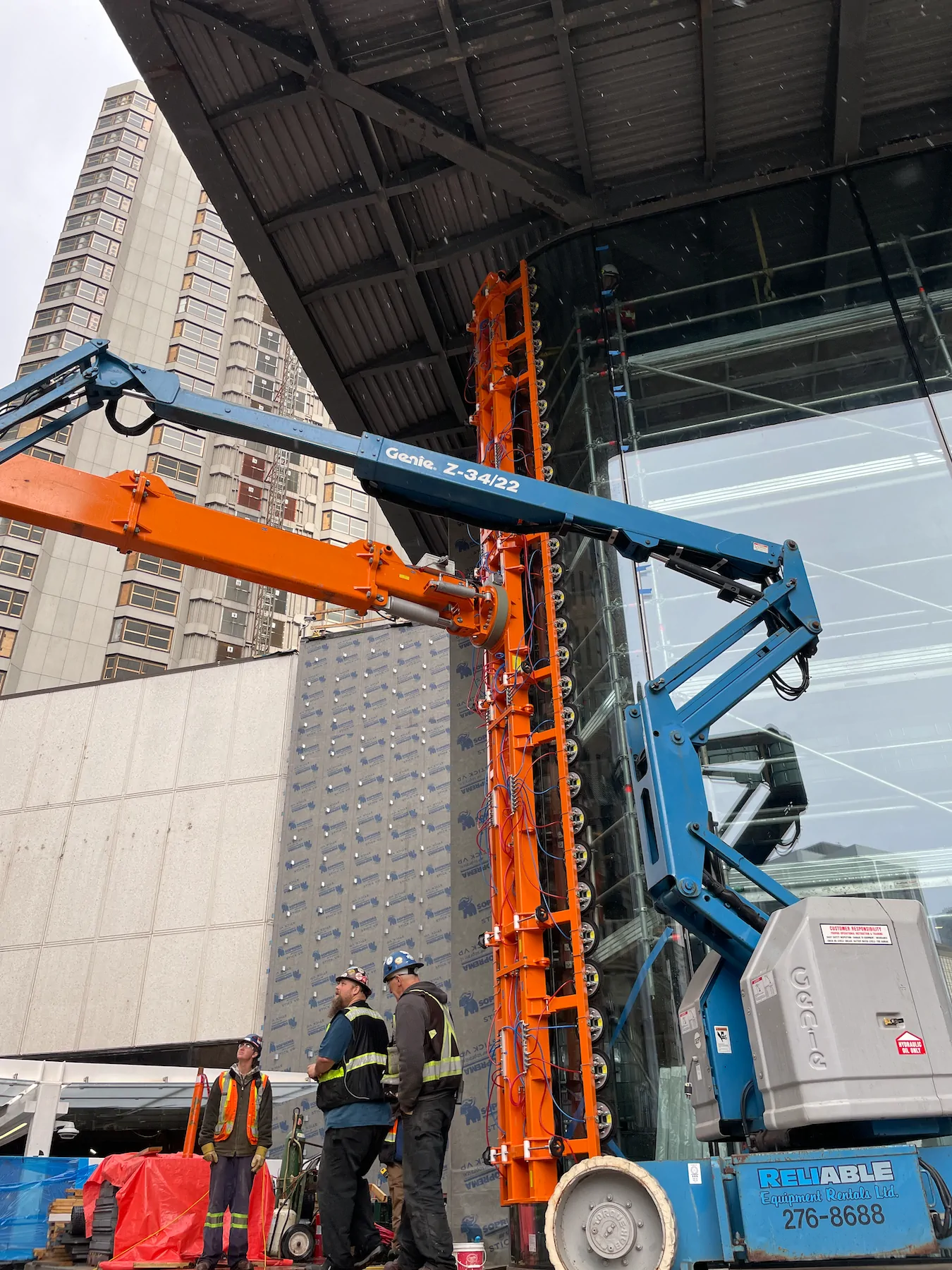 Tall orange pad rail with a row of round pads raised beside a blue boom lift and an orange arm under a steel canopy, with three workers below.