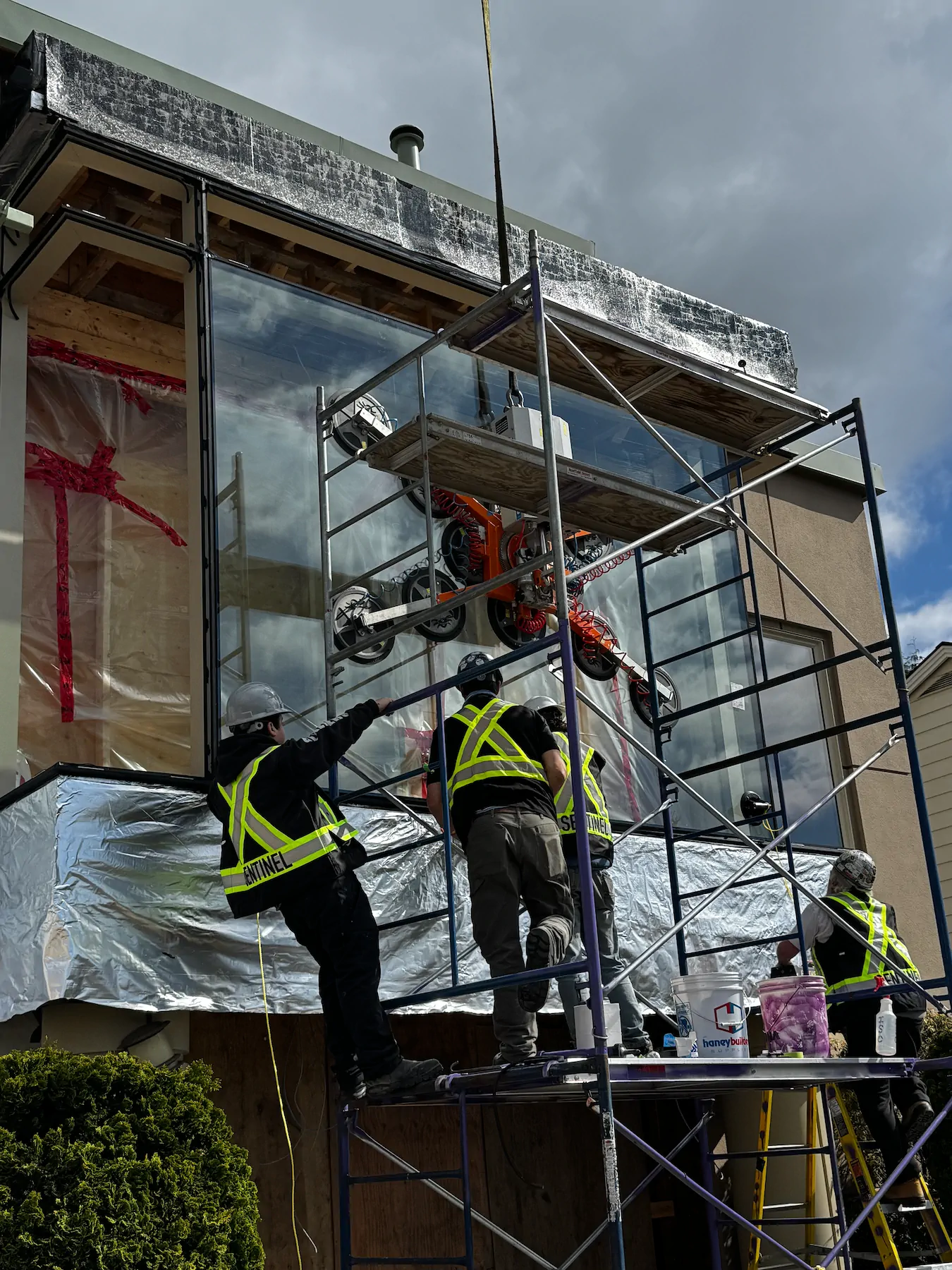 Vacuum lifter holding a window pane at a house facade behind scaffolding while workers on the scaffold guide it.