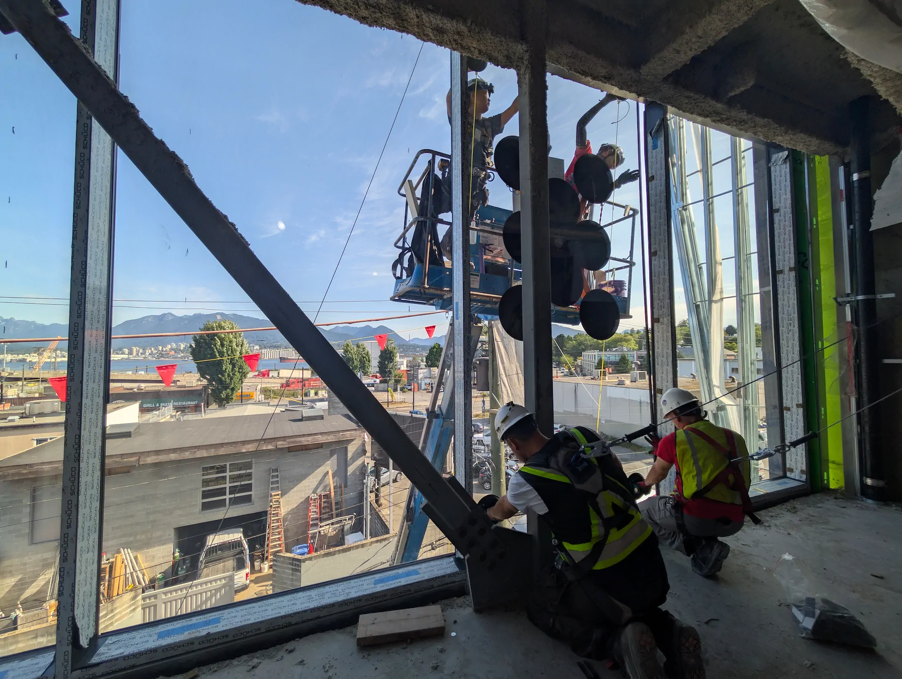 Crew inside a building under construction guiding a glass pane while a vacuum lifter with large pads holds it from a lift platform outside.
