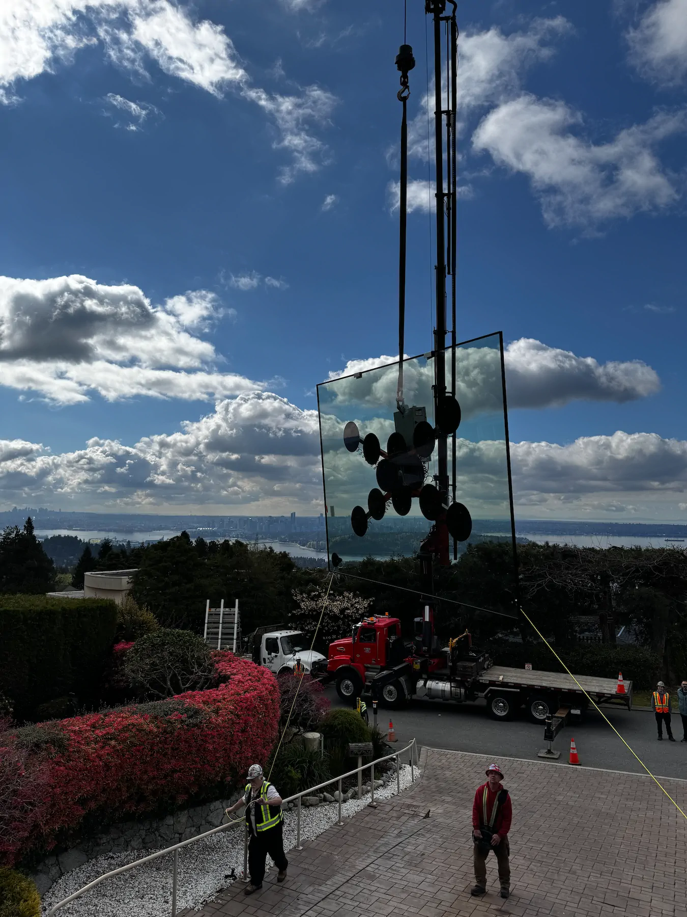 Truck-mounted crane holding a large glass pane on a vacuum lifter above a driveway, with mountains and water in the distance.