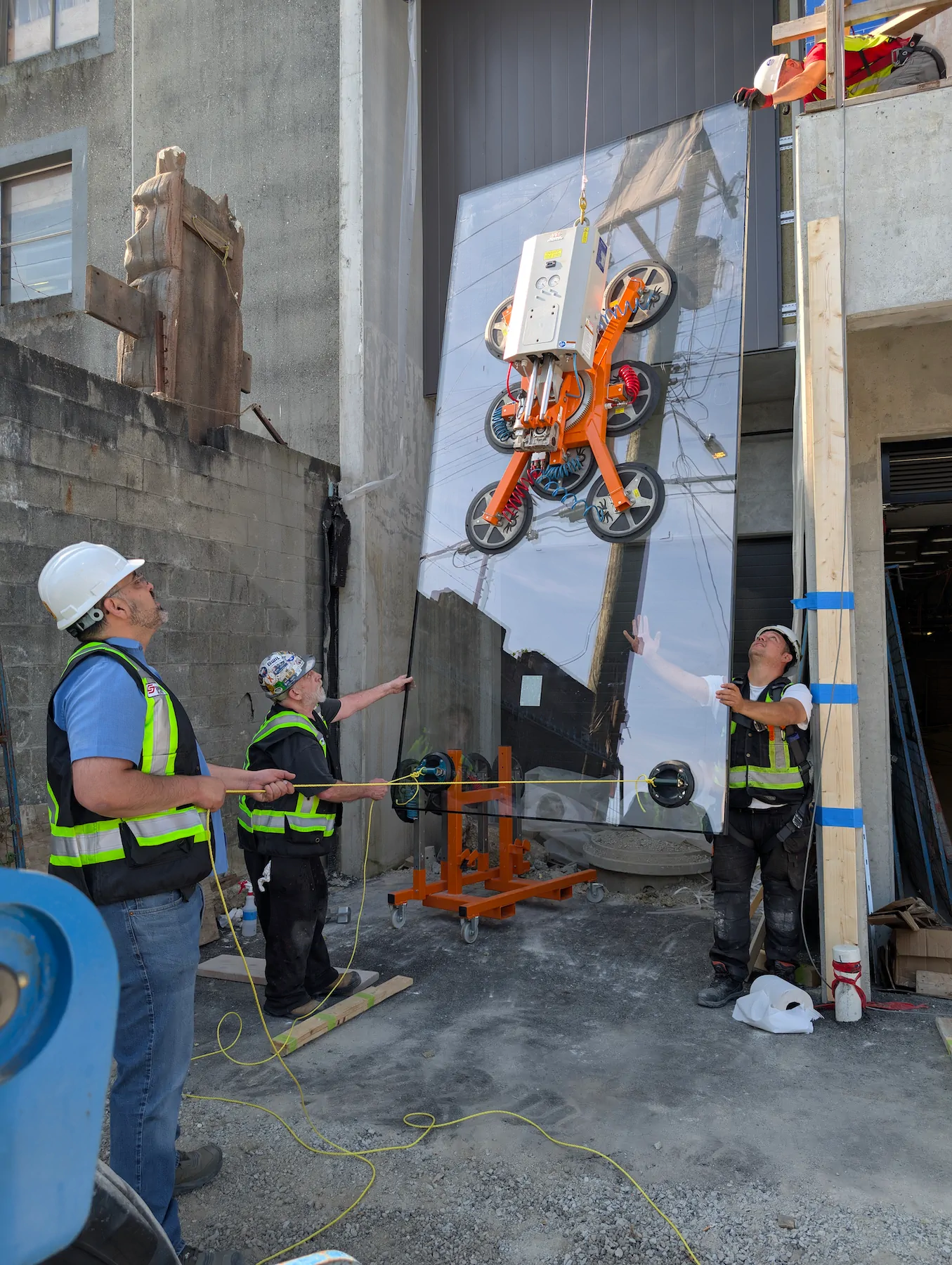 Vacuum lifter in a compact pad layout holding a glass pane beside a concrete wall while three workers guide it.