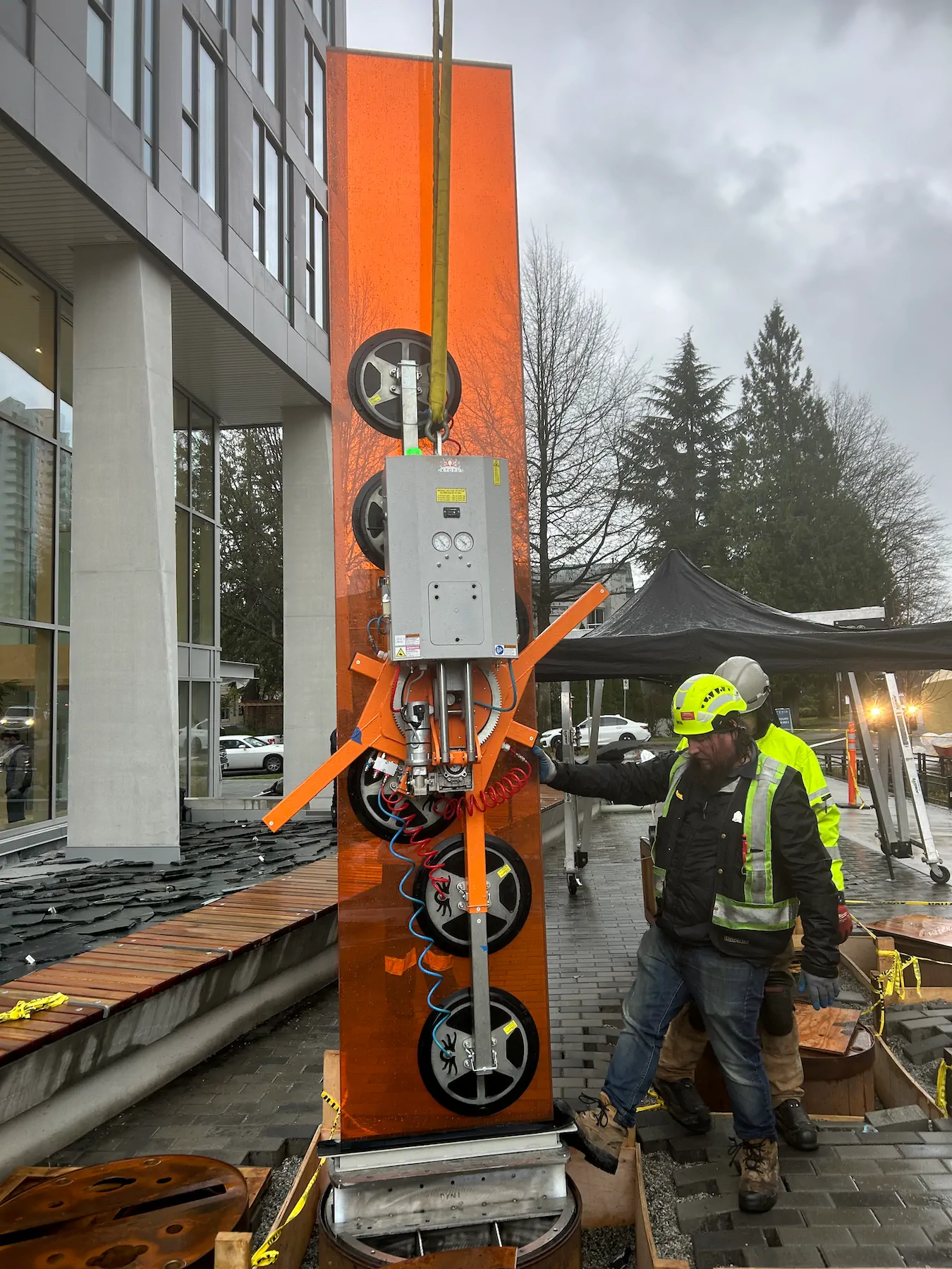 Worker steadying a tall orange tinted glass pane held by a vacuum lifter on a construction site next to a residential tower.