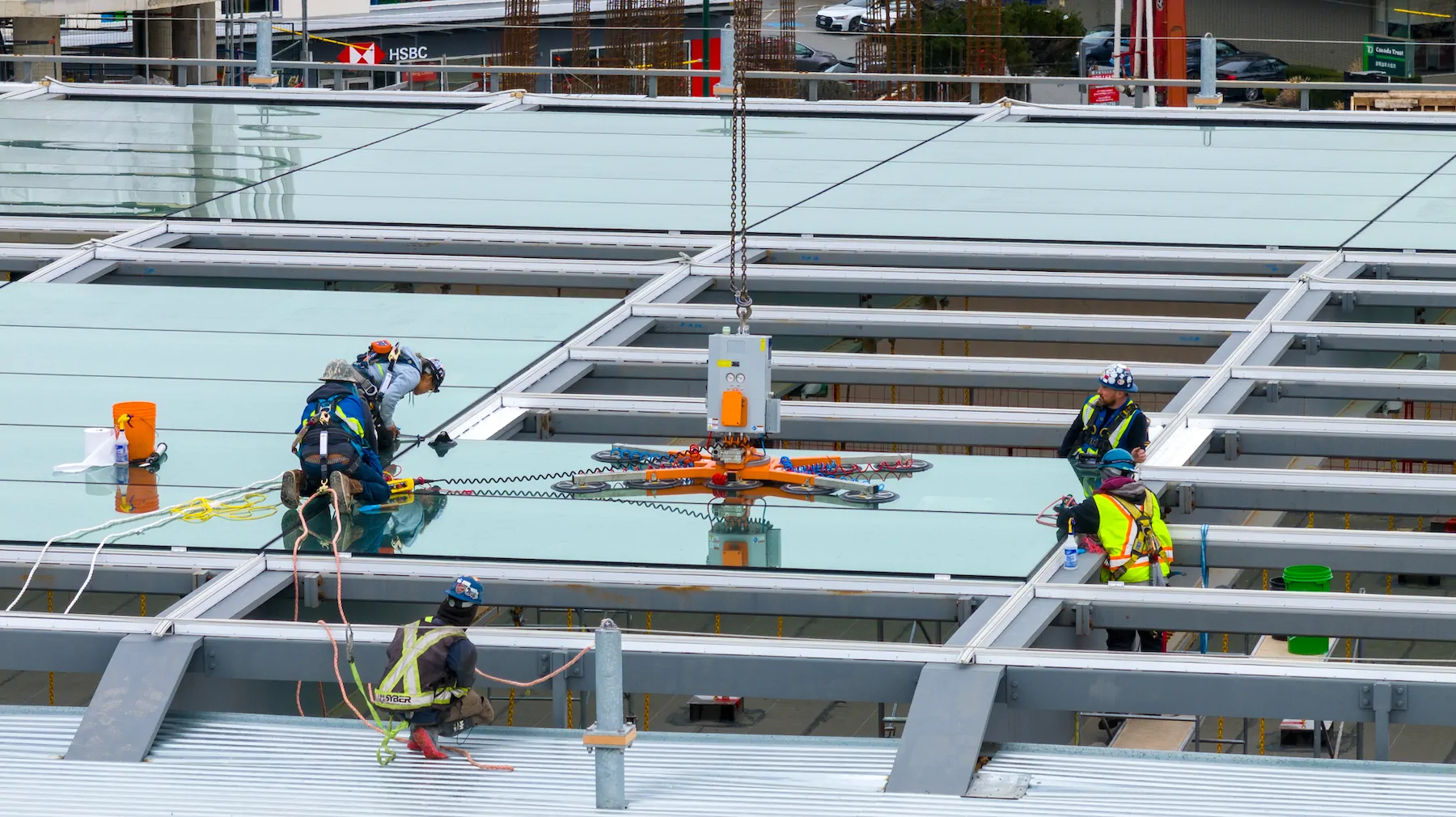 Crew in safety harnesses guiding a vacuum lifter that holds a large glass panel above the steel roof frame of a commercial building.
