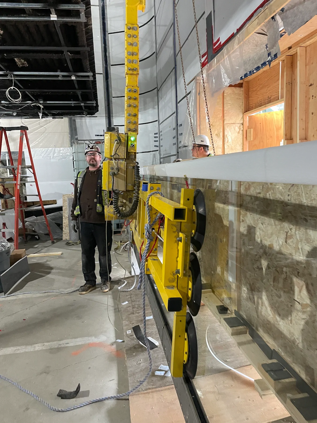 Worker standing beside the yellow vacuum lifter, whose frame and black suction pads hold a large glass pane inside a building under construction.