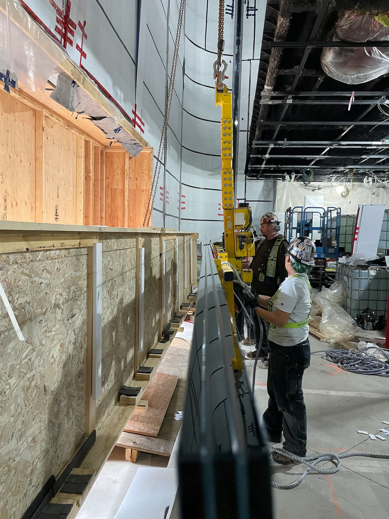 Two workers guiding a long glass pane held by the yellow vacuum lifter, which hangs from a hoist chain inside a building under construction.