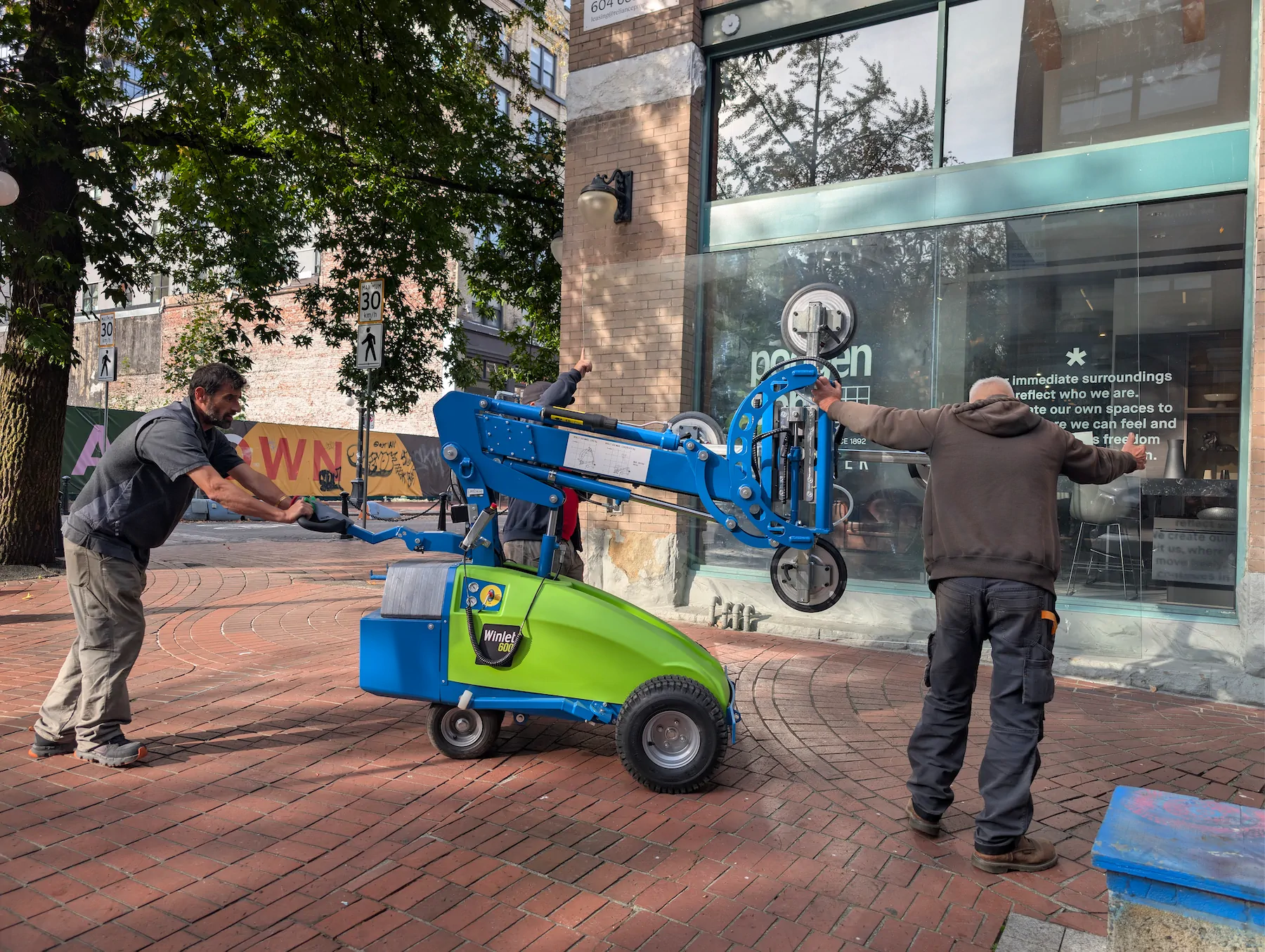 Three workers transporting a large, clear glass pane across a red brick sidewalk toward a commercial building using a blue and green Winlet 600 robot.