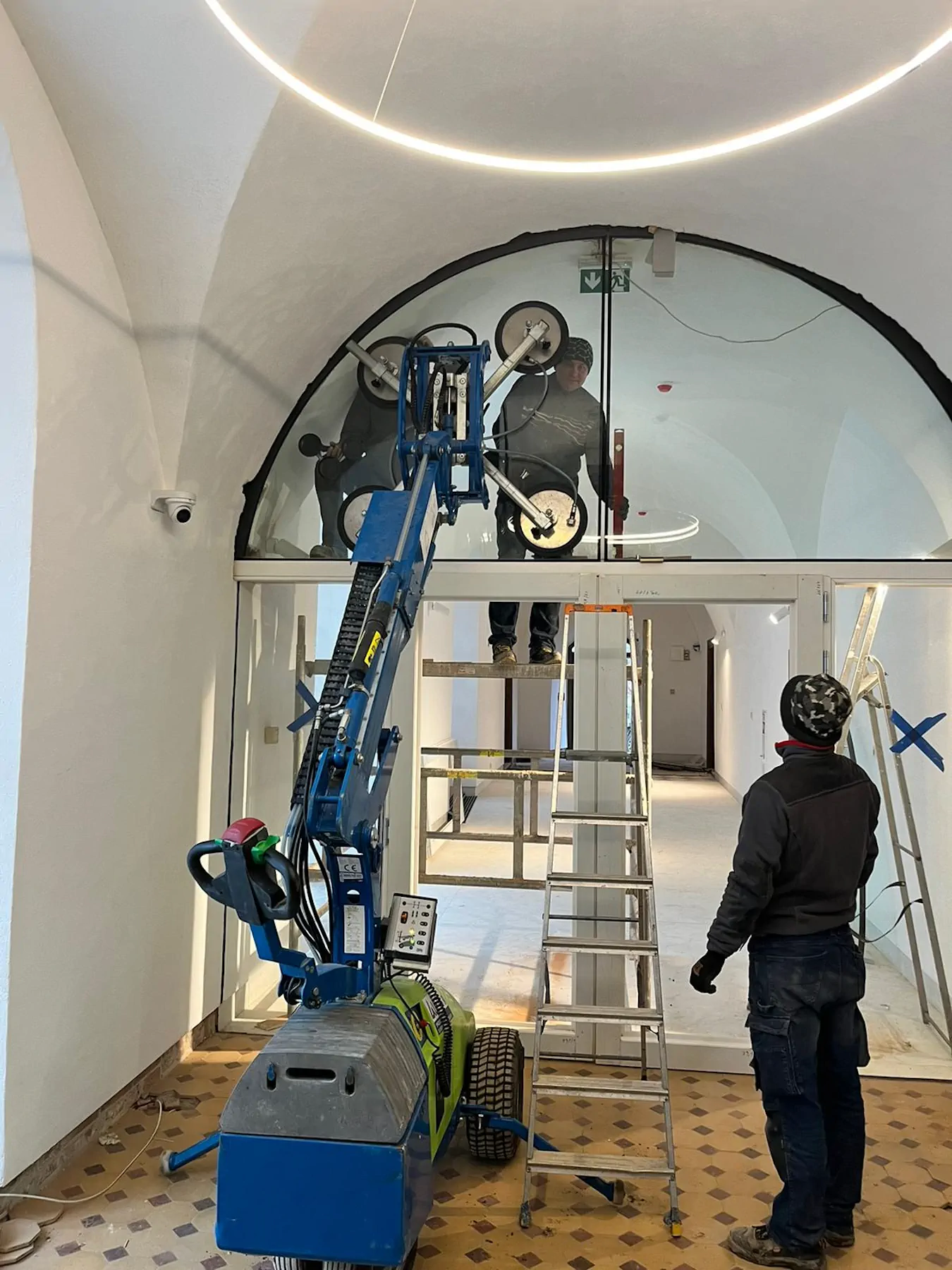 A Winlet 600 robot lifting a large arched glass pane above indoor double doors while three workers guide it into place using a ladder and scaffolding.
