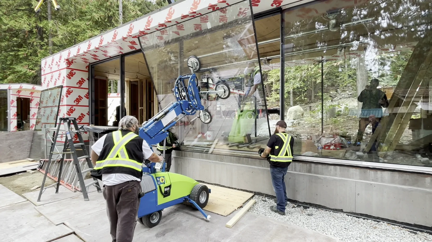 Two construction workers using a blue and green Winlet 600 glazing robot to lift a large glass window pane into a house frame on a wooded construction site.