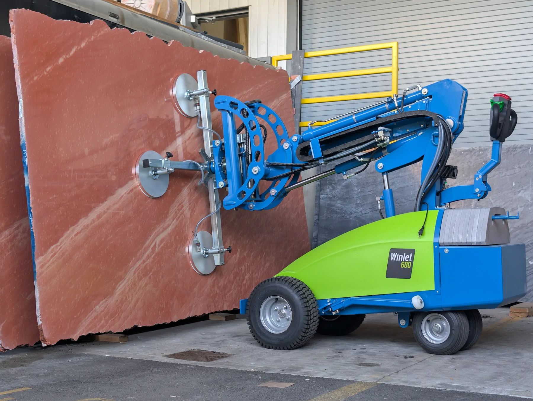 A blue and green Winlet 600 robot using four vacuum suction cups to hold a large, heavy reddish-brown stone slab vertically on a concrete floor.