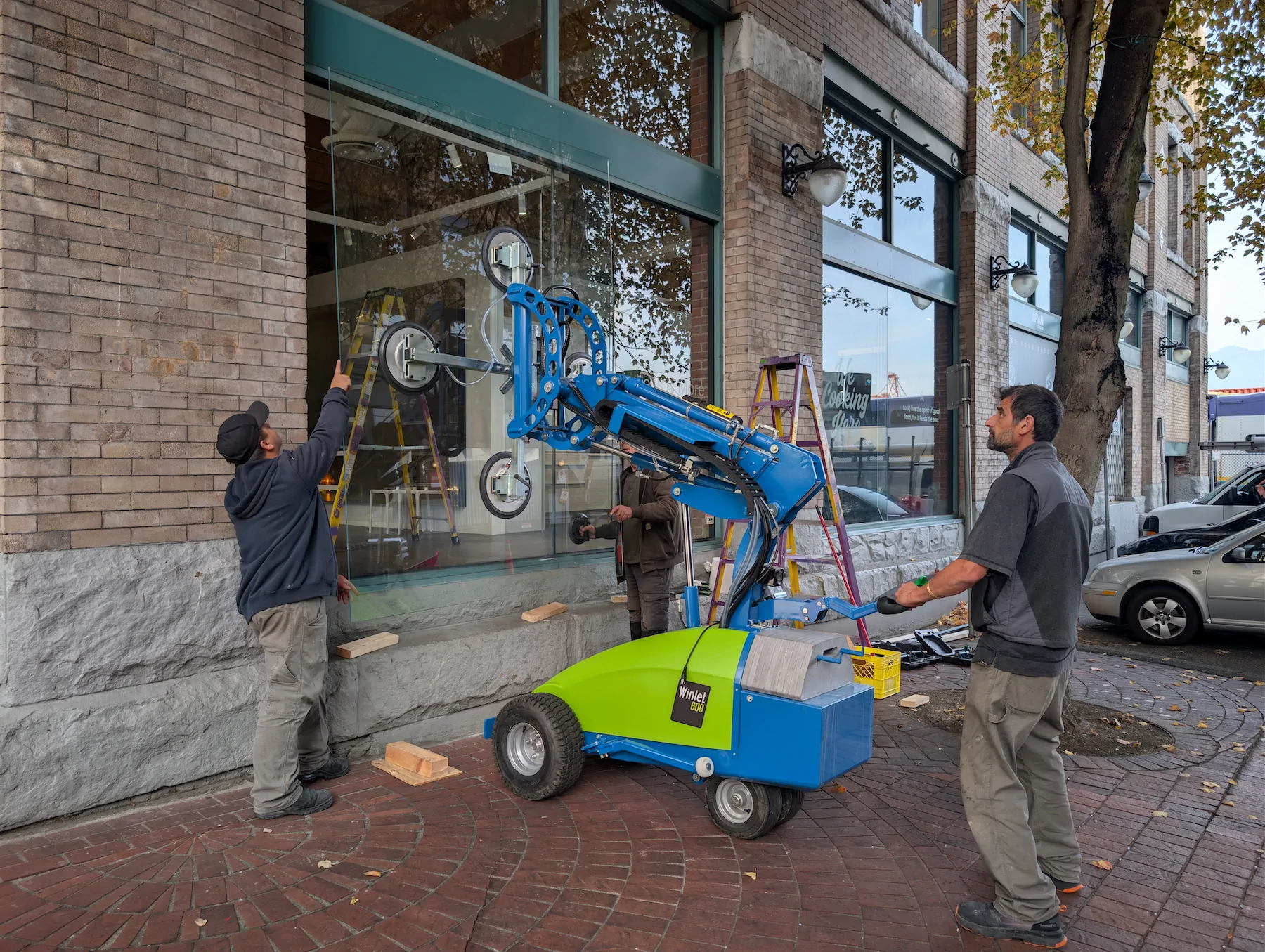 Three workers using a Winlet 600 robot to position and install a large glass pane into a brick commercial storefront on a brick sidewalk.