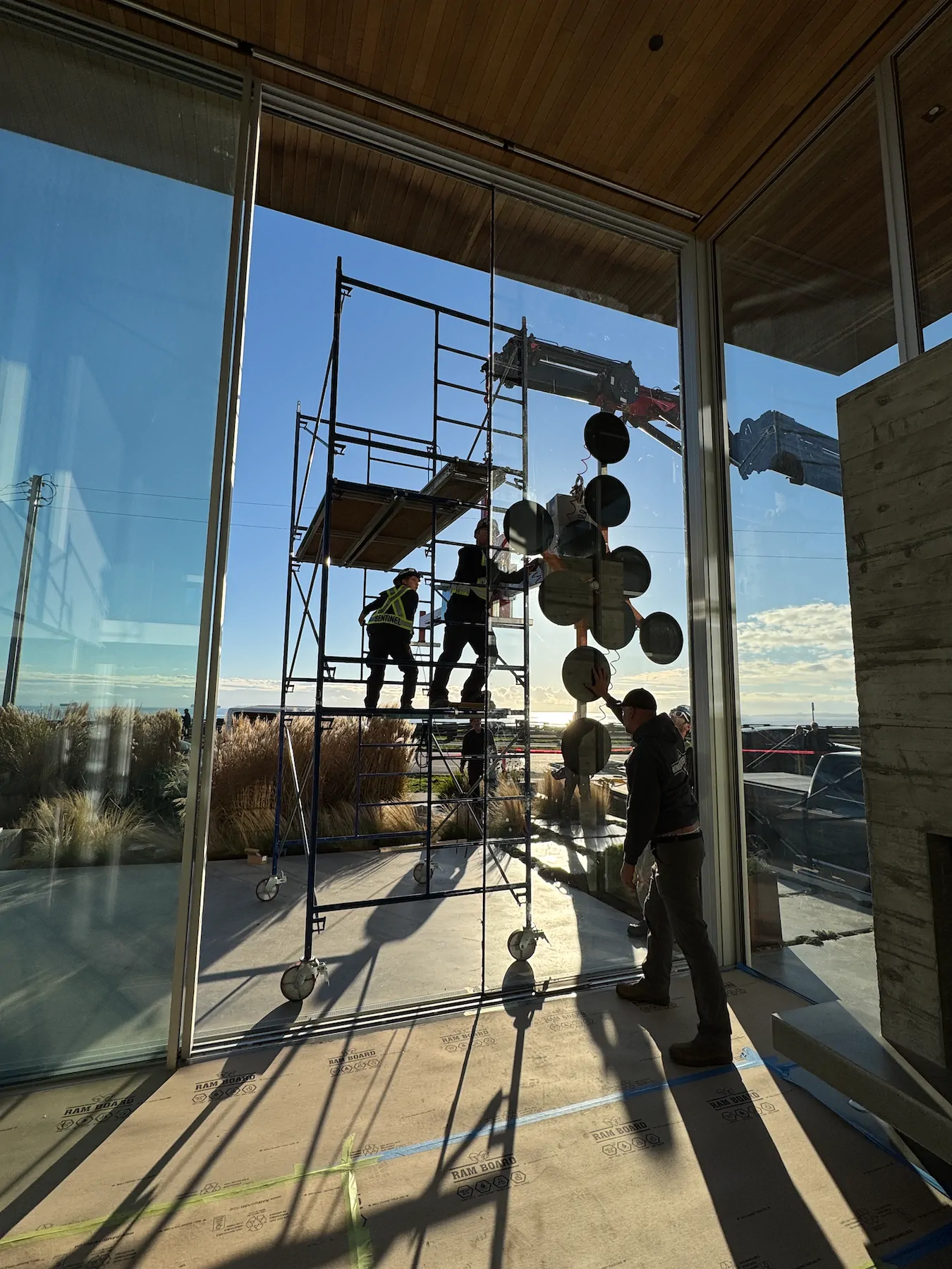 Vacuum lifter with round pads holding a tall glass pane in a glazed opening while workers on a scaffold tower guide it.
