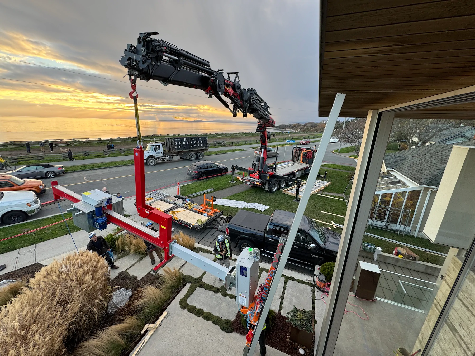 View from above of a truck crane lowering a counterbalance beam beside a house, with a flatbed truck and the water in the background.
