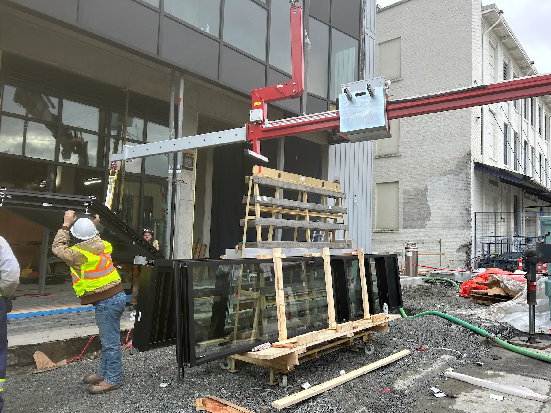 Counterbalance beam suspended above a storefront site while a worker steadies a glass unit beside a wooden delivery rack.
