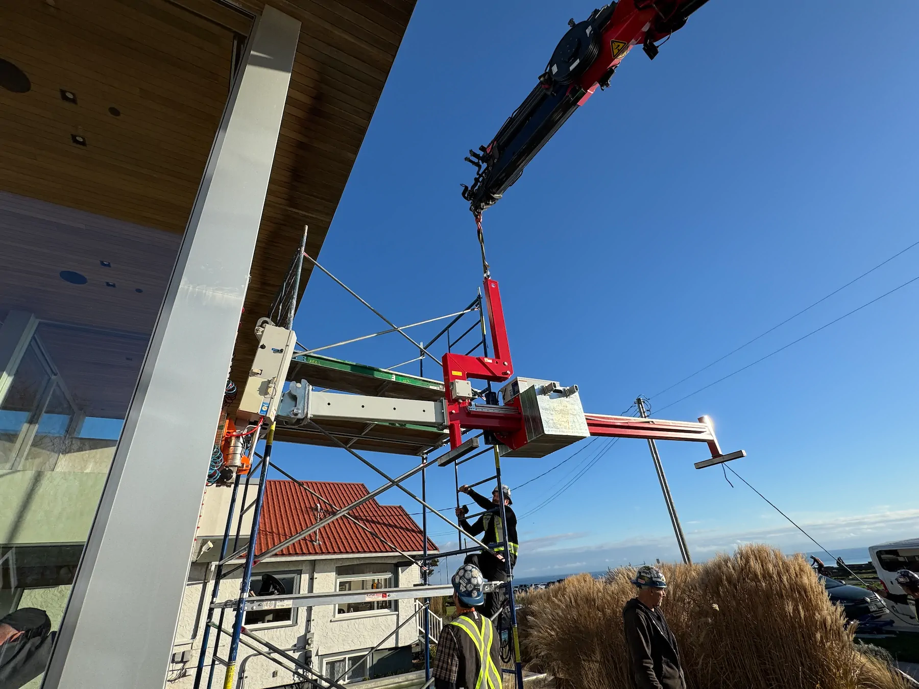 Red counterbalance beam hanging from a truck crane with its forward arm extending beneath a timber roof overhang, and crew on scaffolding below.