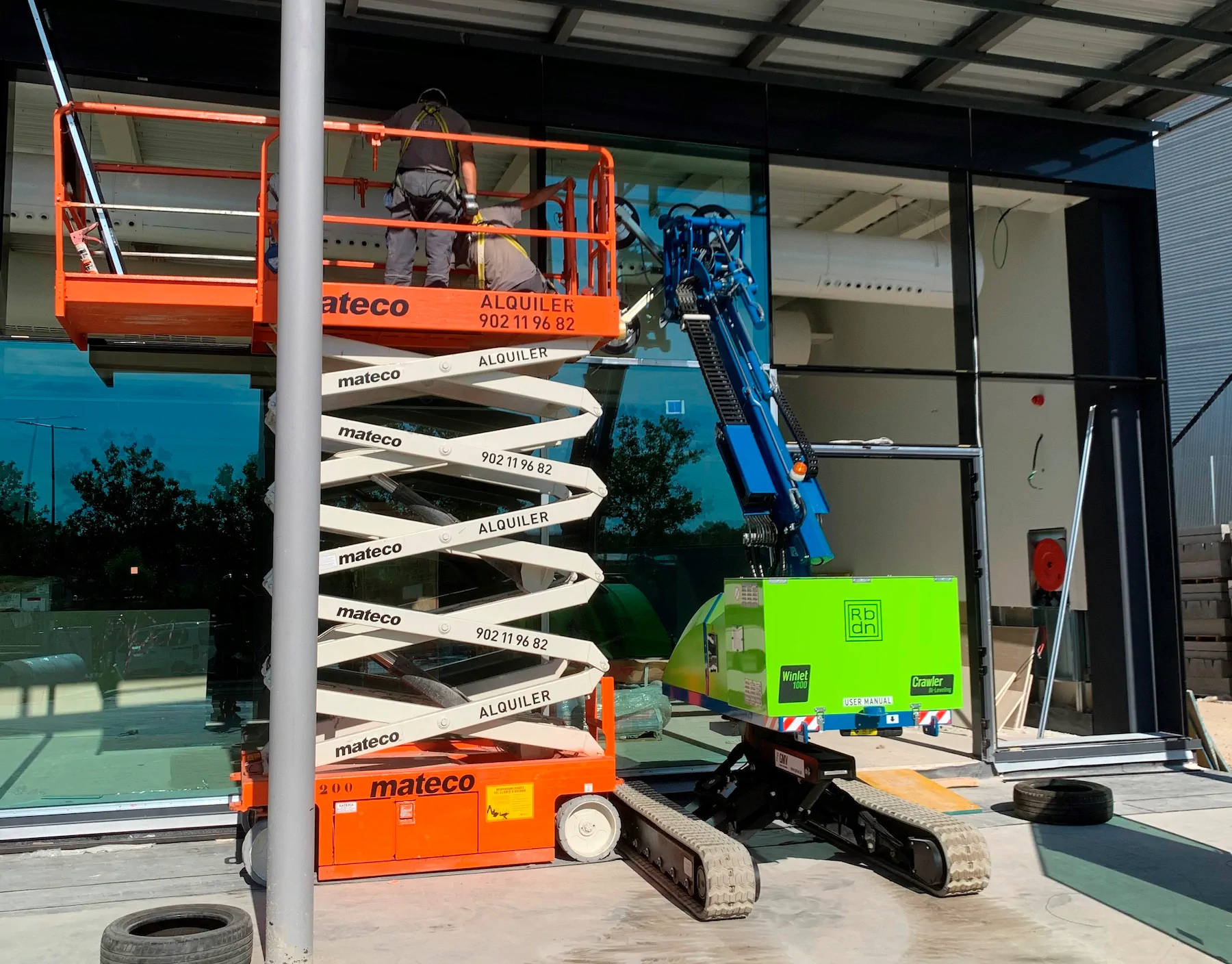 Winlet 1000 Crawler ER next to an orange scissor lift at a glazed building front, its arm raised against the glass.