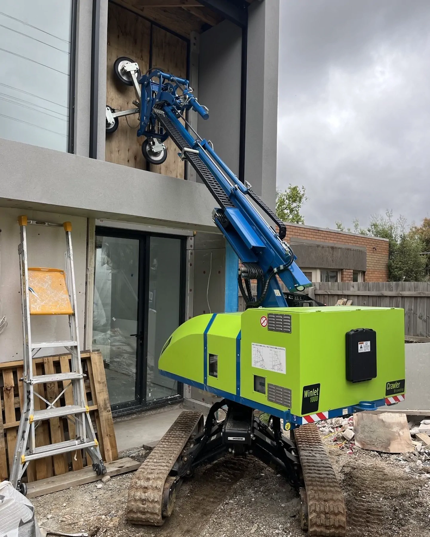 Winlet 1000 Crawler ER on rubble-covered ground with its suction cups against the wall of a house, a ladder and pallets beside it.