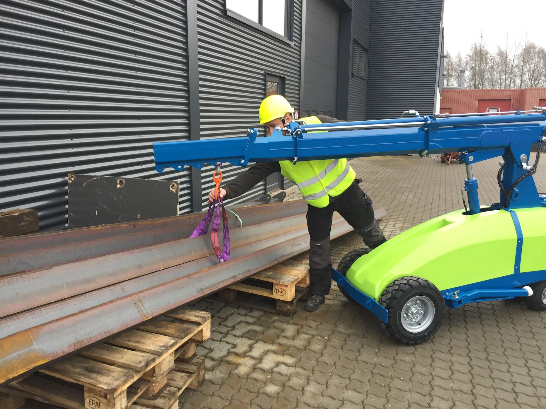 Worker in a hard hat and high-visibility vest guiding steel sections on a pallet while a mini crane holds them from its boom hook.