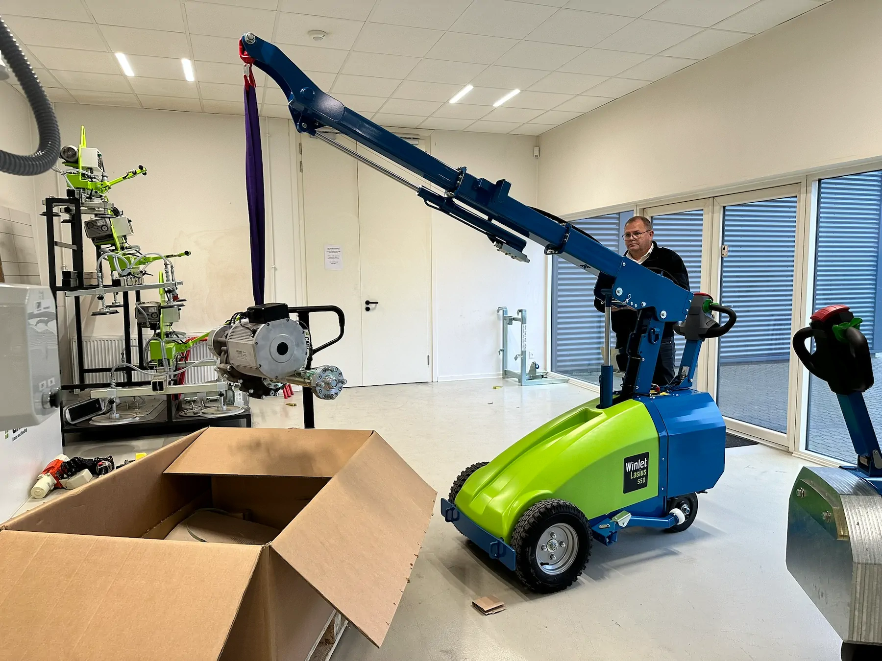 Mini crane working inside a workshop, lifting a load on a purple sling while an operator stands at the controls.