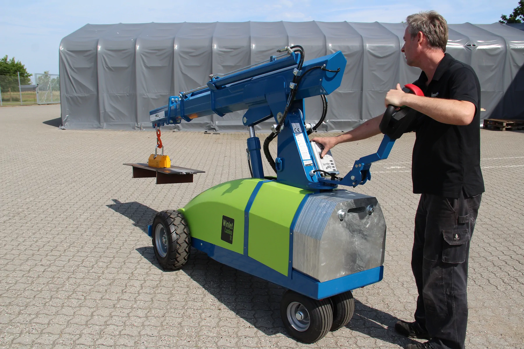 Operator walking behind a wheeled mini crane that carries a steel plate hanging from a lifting magnet on a paved yard.