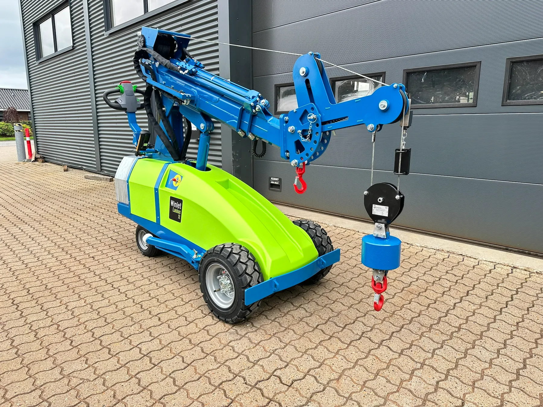 Wheeled mini crane with its boom raised and a winch line running over the boom tip to a hook block, standing beside a grey industrial building.