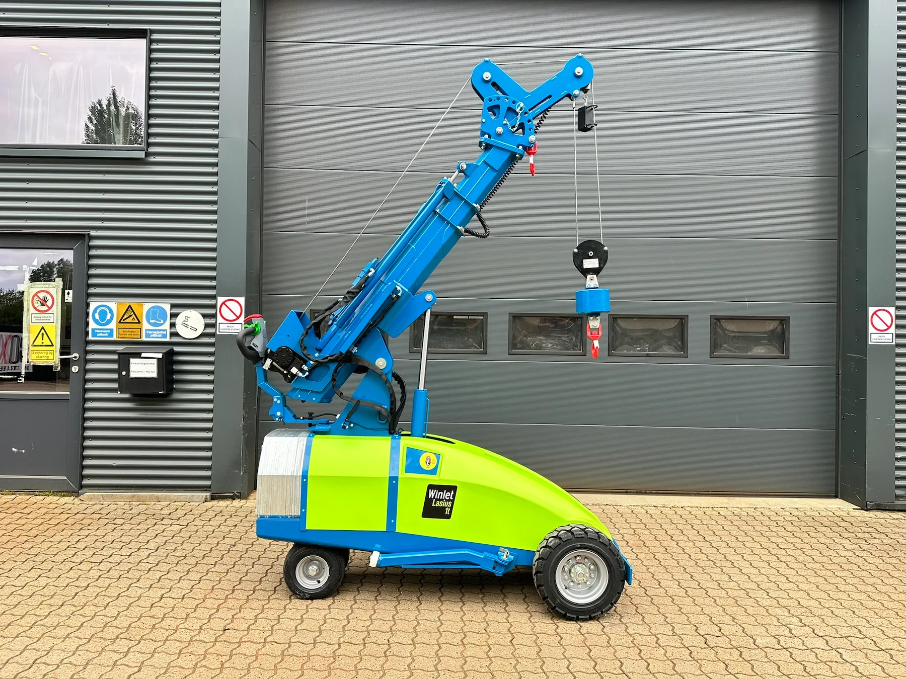 Side view of a wheeled mini crane with its boom raised high and a winch line lowering a hook block against a grey building wall.