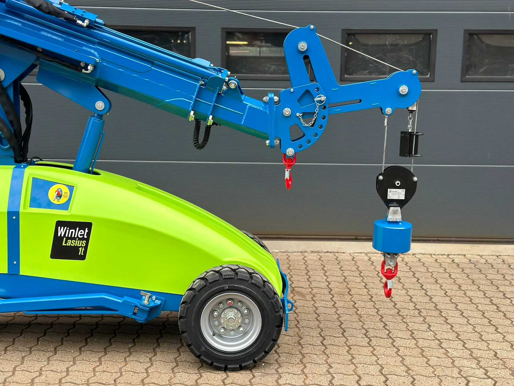 Close view of a wheeled mini crane’s green body, raised blue boom, winch line and hook block against a grey building wall.